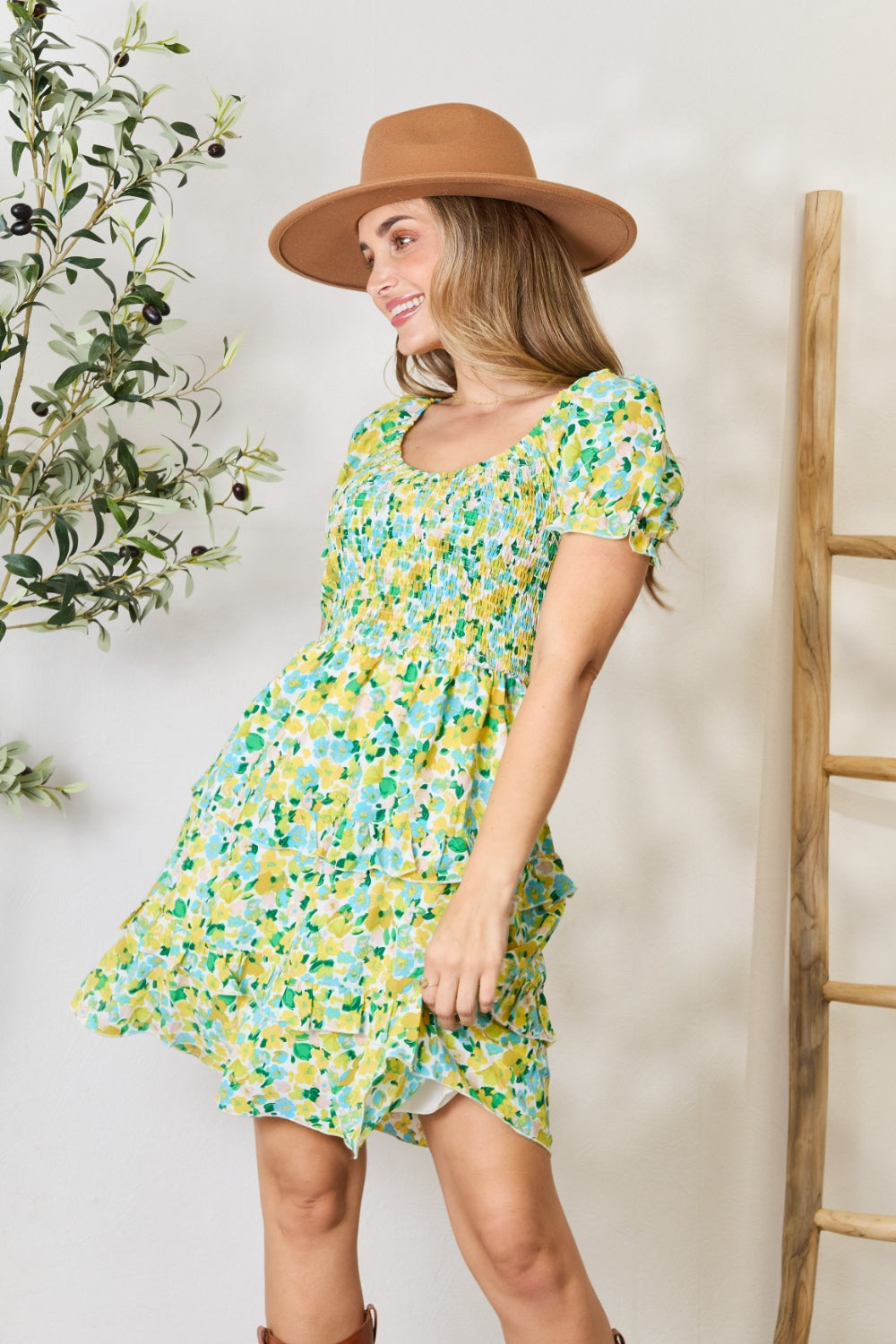 A woman wearing the Devine Smocked Scoop Neck Flounce Sleeve Mini Dress and a brown hat smiles and poses near greenery indoors.