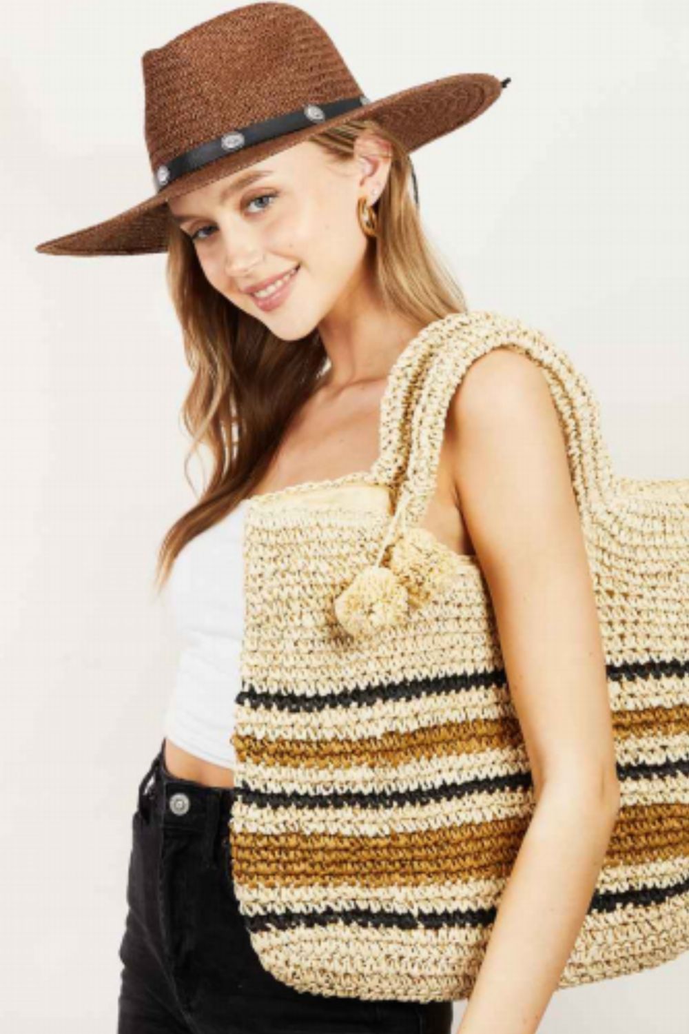 A person wearing a brown hat, white top, and black pants carries the stylish Fame Striped Straw Braided Tote Bag.