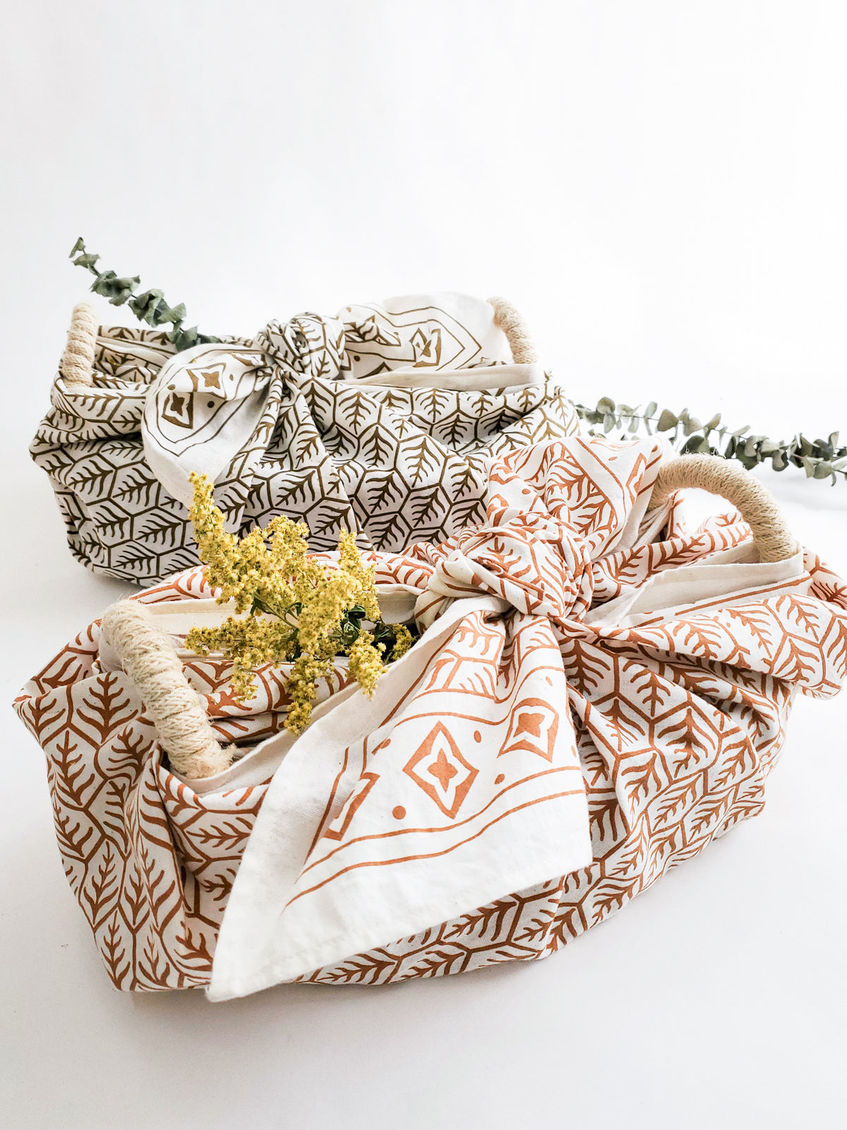 A decorative ceramic plate is beautifully set in a handwoven seagrass basket featuring handles, called the Bread Warmer & Basket Gift Set with Tea Towel - Flower. Below the basket are two patterned towels, crafted by Fair Trade artisans, showcasing leaf designs in black and orange.