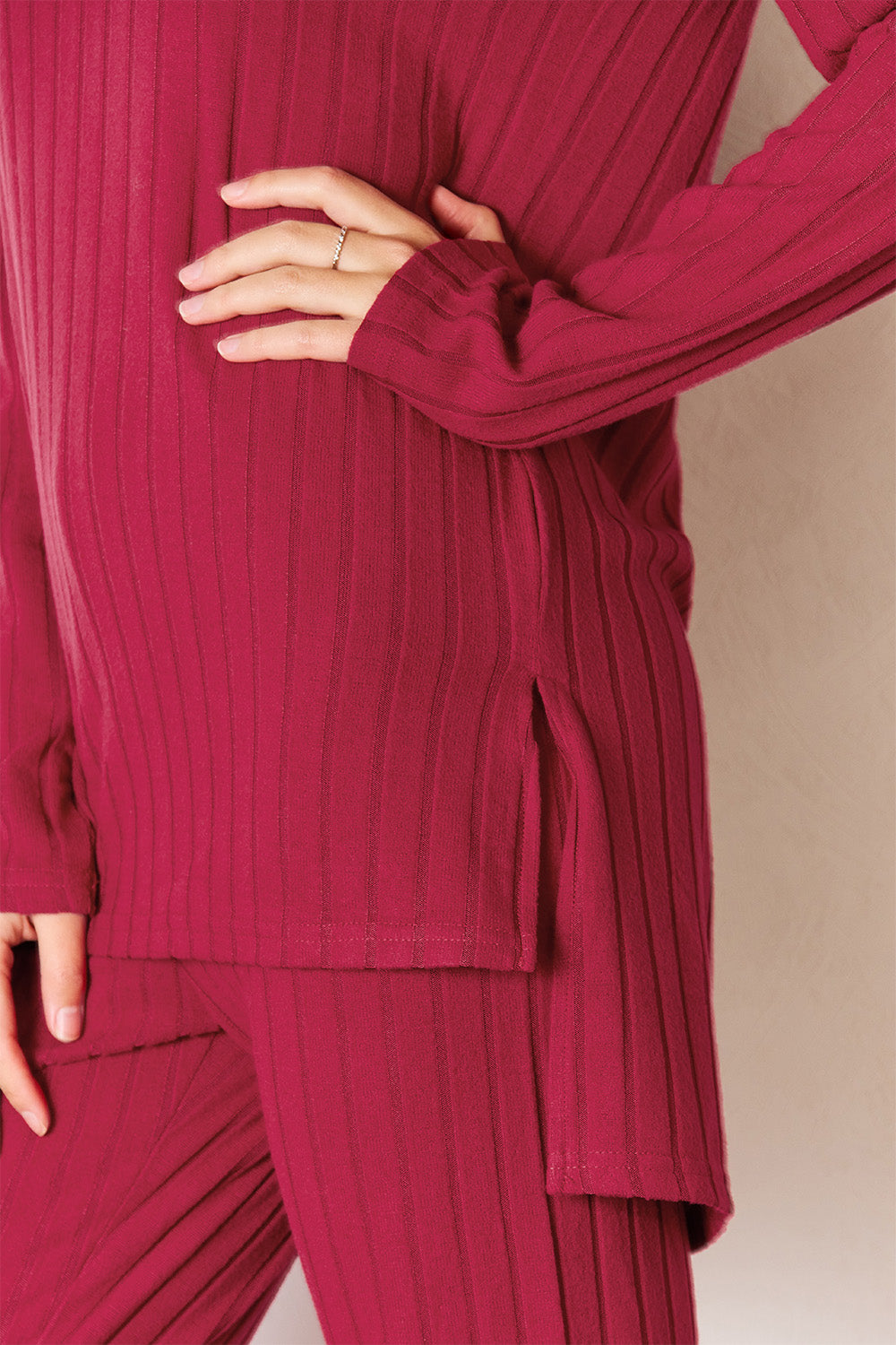 A woman in a Basic Bae Full Size Ribbed Round Neck High-Low Slit Top and Pants Set poses indoors near a wooden ladder. She is wearing the matching maroon slightly stretchy top and pants, paired with beige heels. Pampas grass in a white vase is visible in the foreground. For care, machine wash cold.