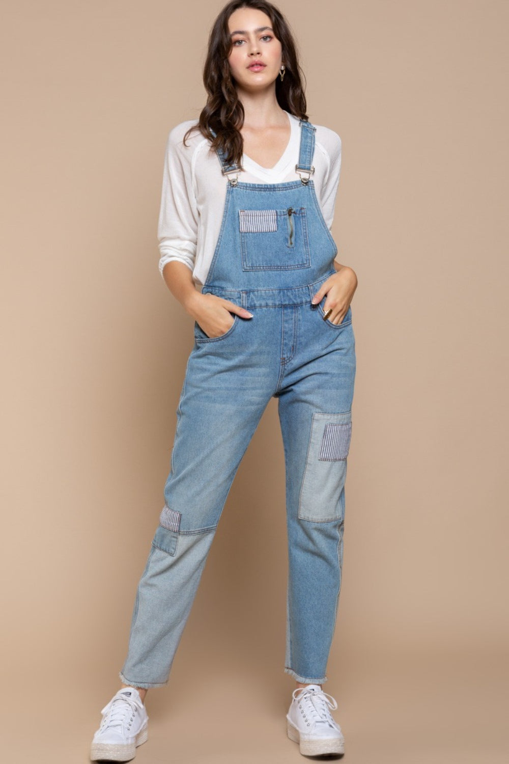 A woman kneels on one knee against a beige background, wearing a beige hat and white long-sleeve shirt, paired with POL Front Chest Zipper Slim Leg Denim Overalls featuring patches.