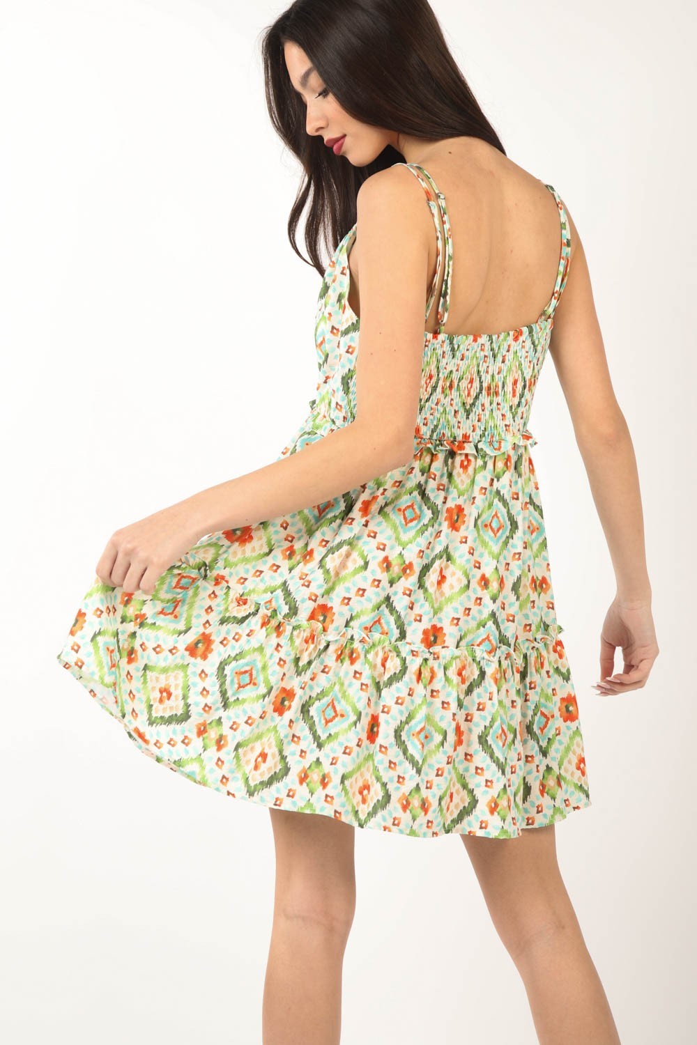 A woman in a VERY J Floral Back Smocked Ruffled Mini Dress poses against a white background. She holds the hem with both hands, and her dark hair is worn loose.