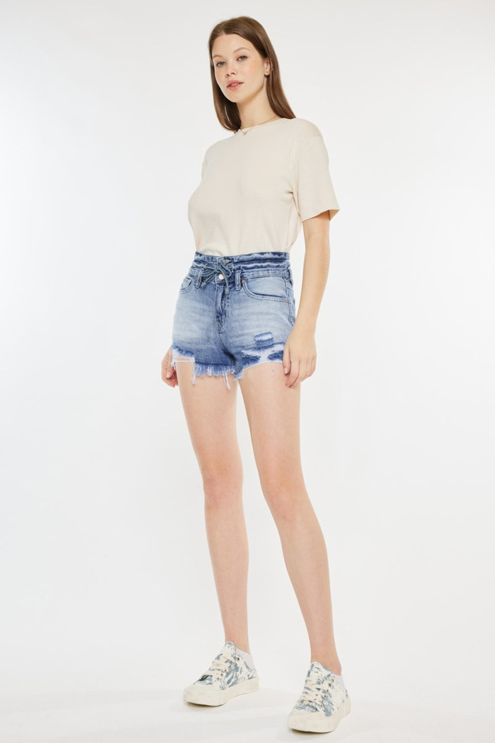 A person wearing a beige t-shirt and Kancan High Rise Frayed Hem Denim Shorts, paired with patterned sneakers, stands against a white background—an ideal addition to any summer wardrobe.