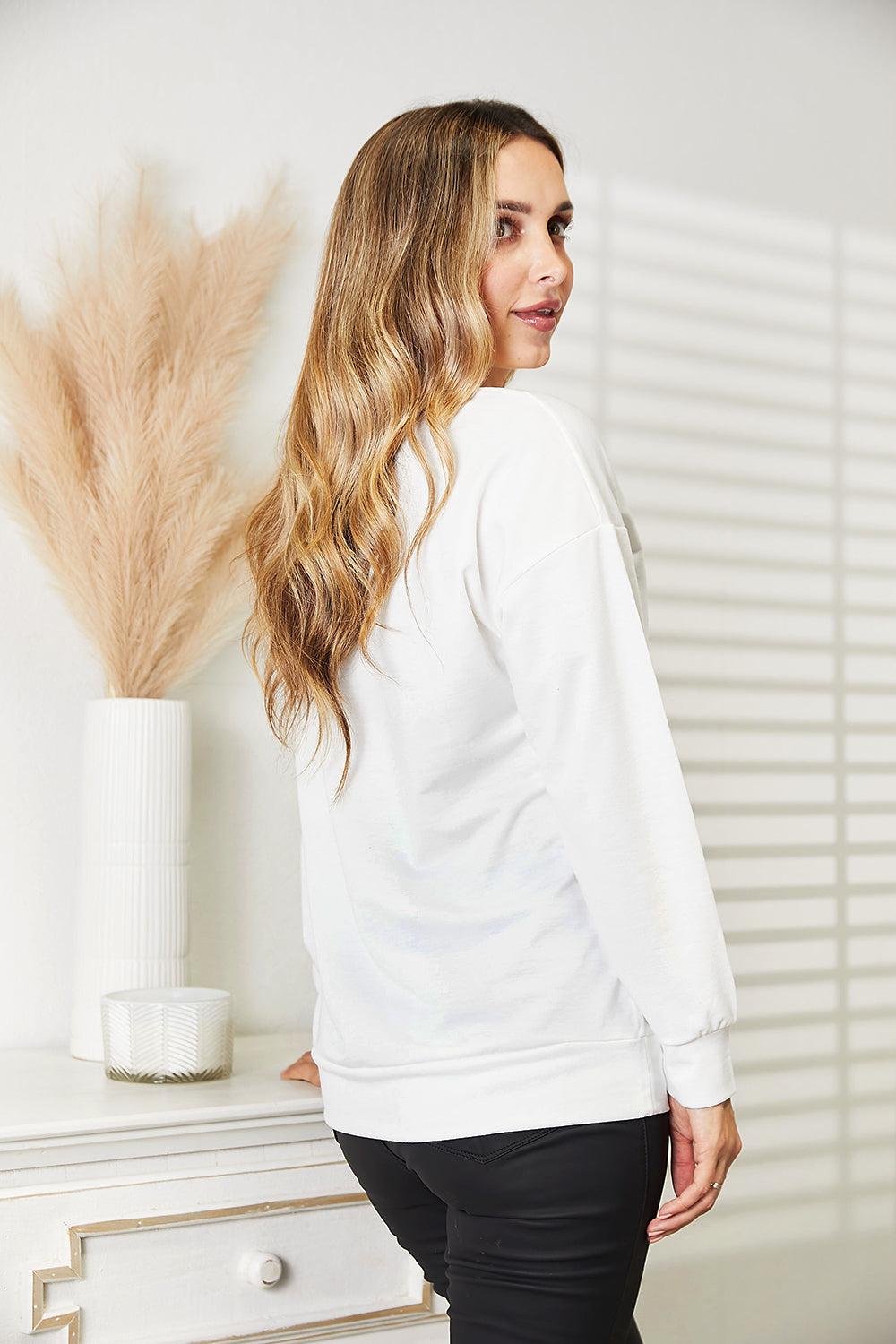 A woman stands indoors wearing a trendy and comfortable outfit—a white Simply Love Graphic Dropped Shoulder Round Neck Sweatshirt, featuring a dripping lip design, paired with black pants. One hand rests on a piece of furniture.