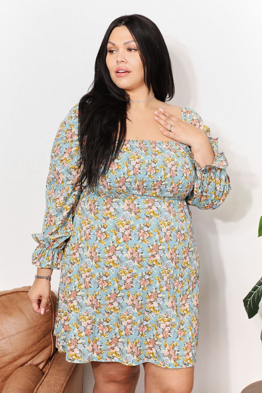 A woman in the Honey Floral Smocked Flounce Sleeve Square Neck Dress stands indoors, holding the hem of her dress. She is smiling slightly with her eyes looking downward. Nearby, a plant and a brown couch complete the cozy scene.