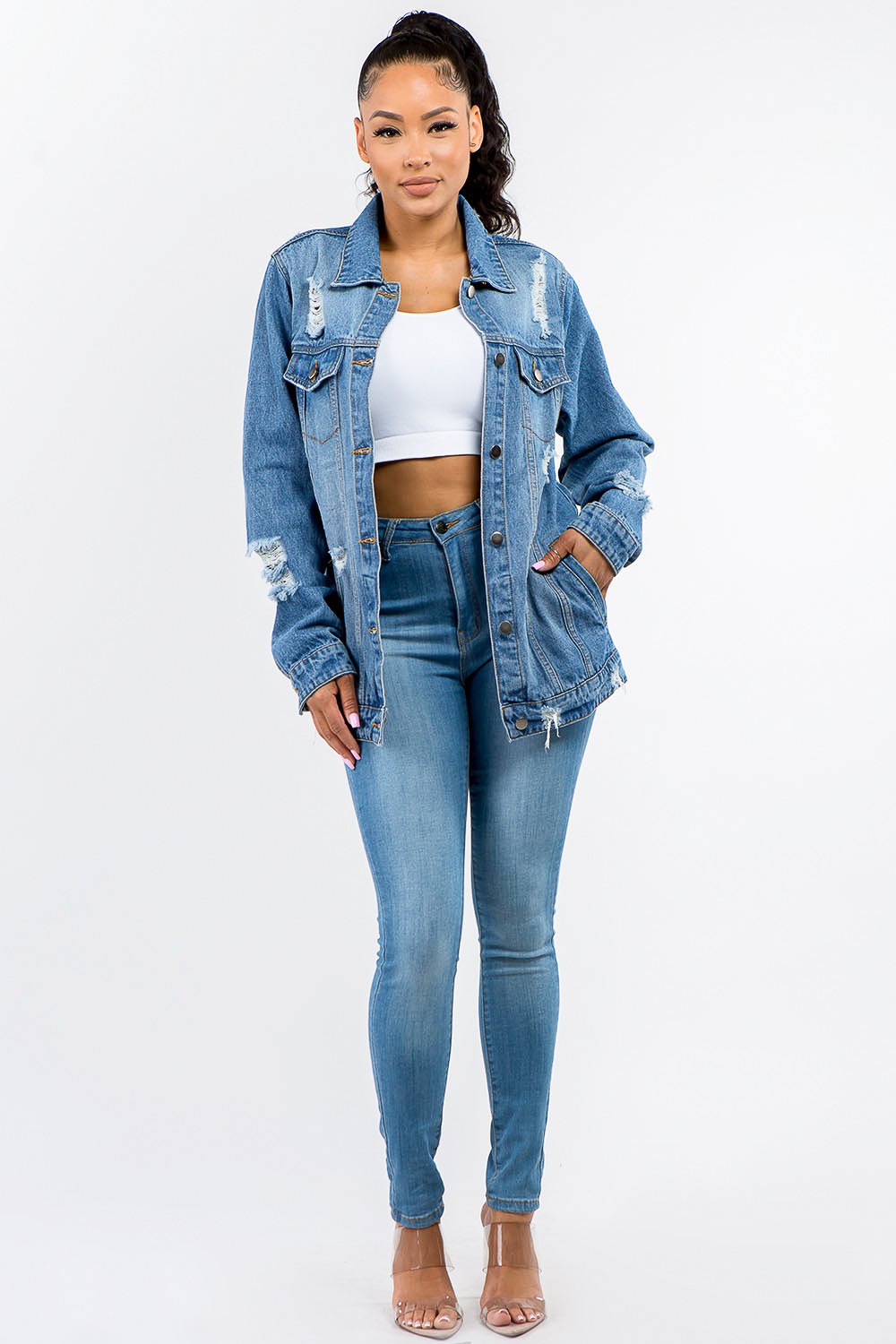 A woman in an American Bazi Full Size Button Up Distressed Denim Jacket with a vintage look stands against a plain background, pairing it perfectly with a white crop top and denim jeans.