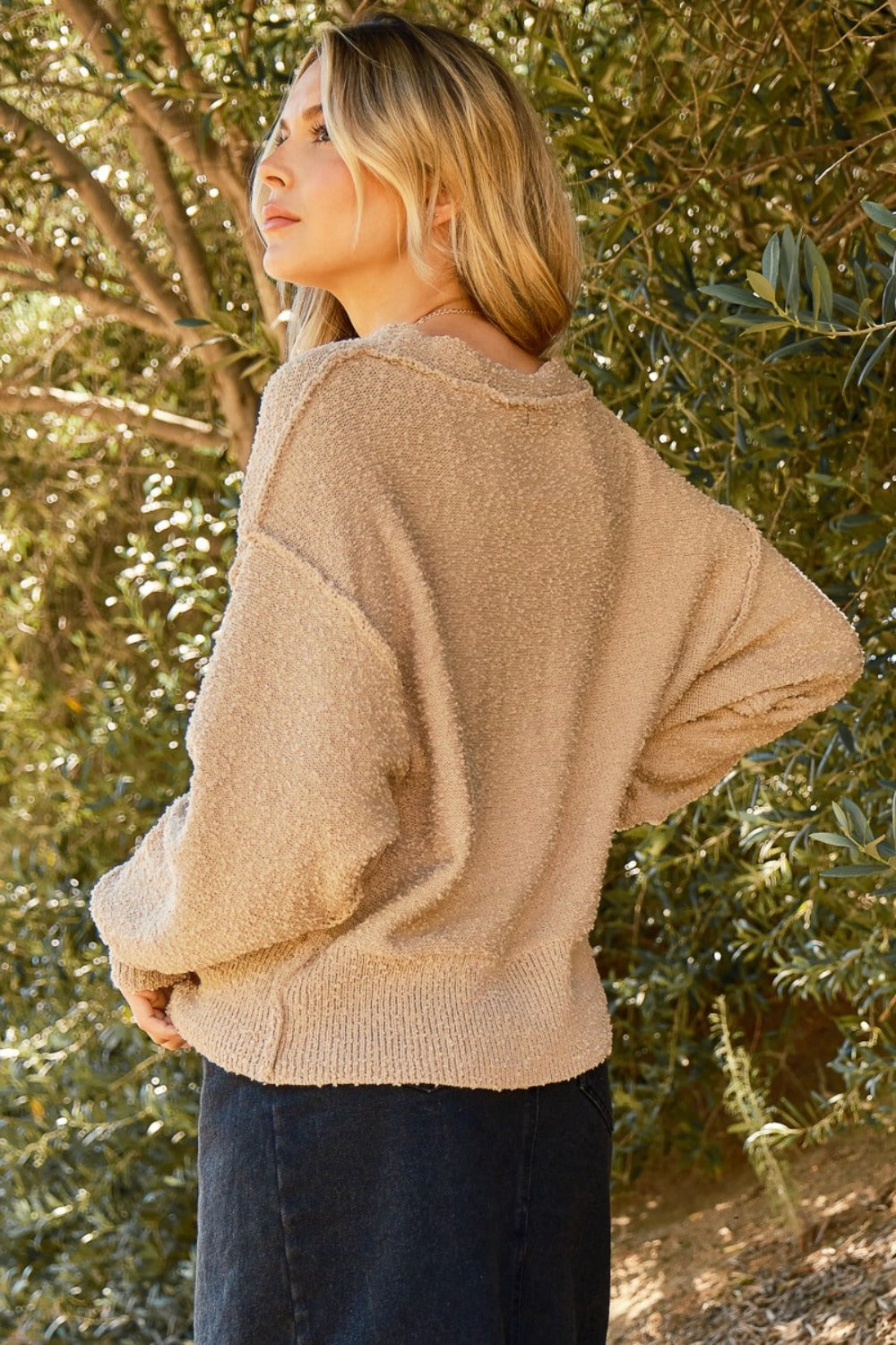 A person stands outdoors wearing the unique And The Why Button Down Popcorn Sweater Cardigan in beige over a white top, looking into the distance. Trees and a mountainous landscape form the serene background.