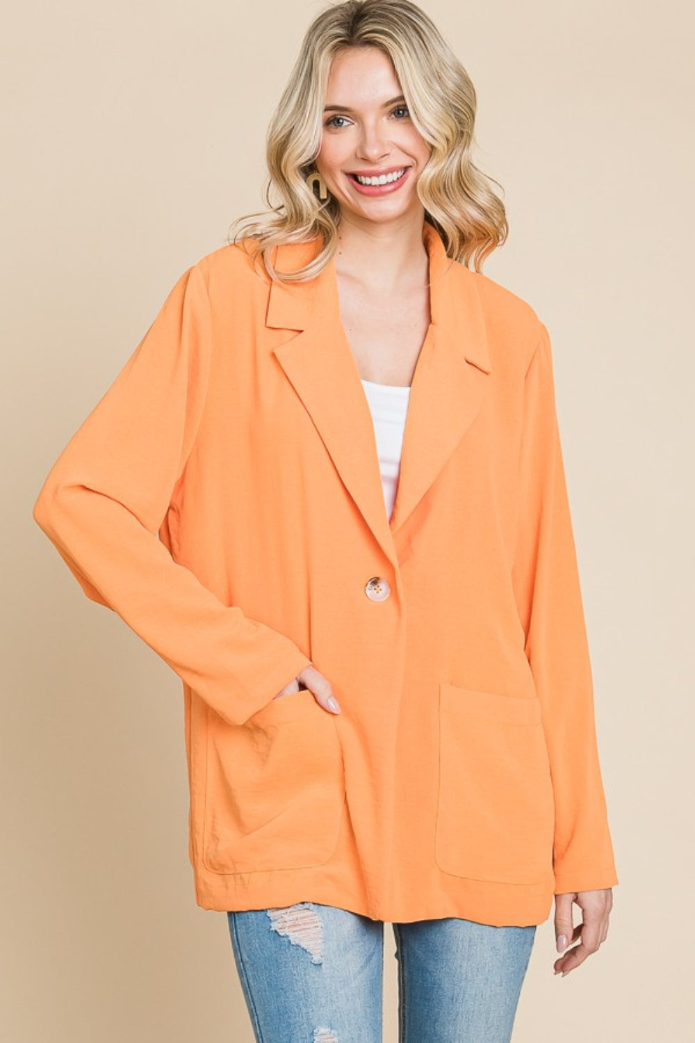 A smiling person models the Culture Code One Button Long Sleeve Blazer with Pockets, a tailored fit in vibrant orange, over a white top and blue jeans. They pose with one hand in their pocket against a plain background, perfectly capturing versatile fashion in this stylish look.