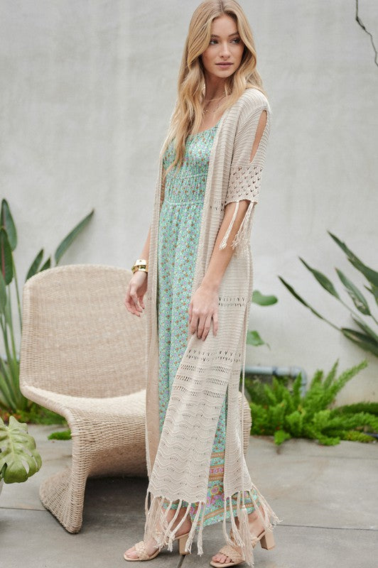 A woman in a green floral dress and a Solid Long Cardigan With Fringe exudes boho-chic vibes as she strolls on a patio, surrounded by outdoor furniture and lush plants.