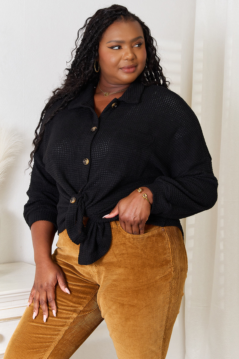 A woman with long hair is wearing a Double Take Waffle-Knit Collared Neck Dropped Shoulder Shirt in black and mustard-colored pants. She stands indoors, resting one hand in her pocket and the other touching her hair.