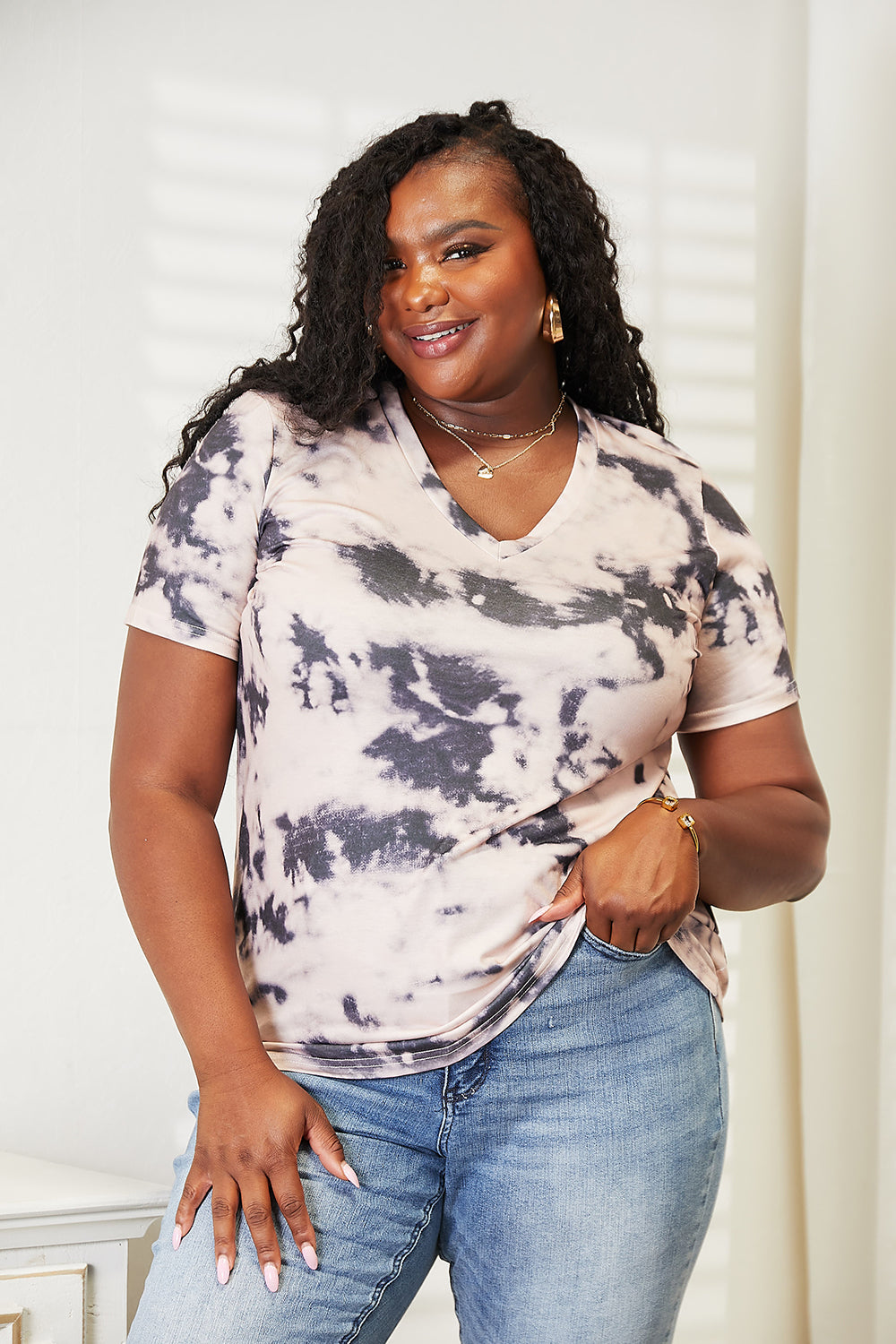 A person in a Double Take Tie-Dye V-Neck T-Shirt and jeans, epitomizing boho-chic, stands indoors, smiling at the camera. A vase with dried plants is visible in the background.