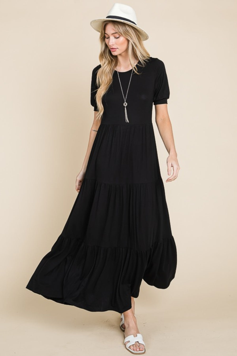 A person wearing the BOMBOM Short Sleeve Tiered Maxi Dress in sleek black stands against a plain beige background, exuding comfort with style. With one hand on their hip and donning white sandals, they effortlessly showcase this versatile piece.