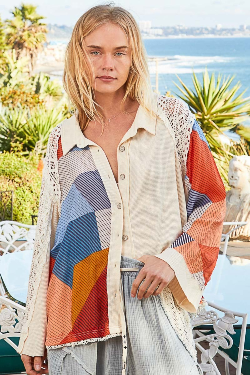 A person stands outdoors in a POL Color Block Crochet Long Sleeve Shirt, exuding a relaxed expression. The coastal background features palm trees and distant buildings.