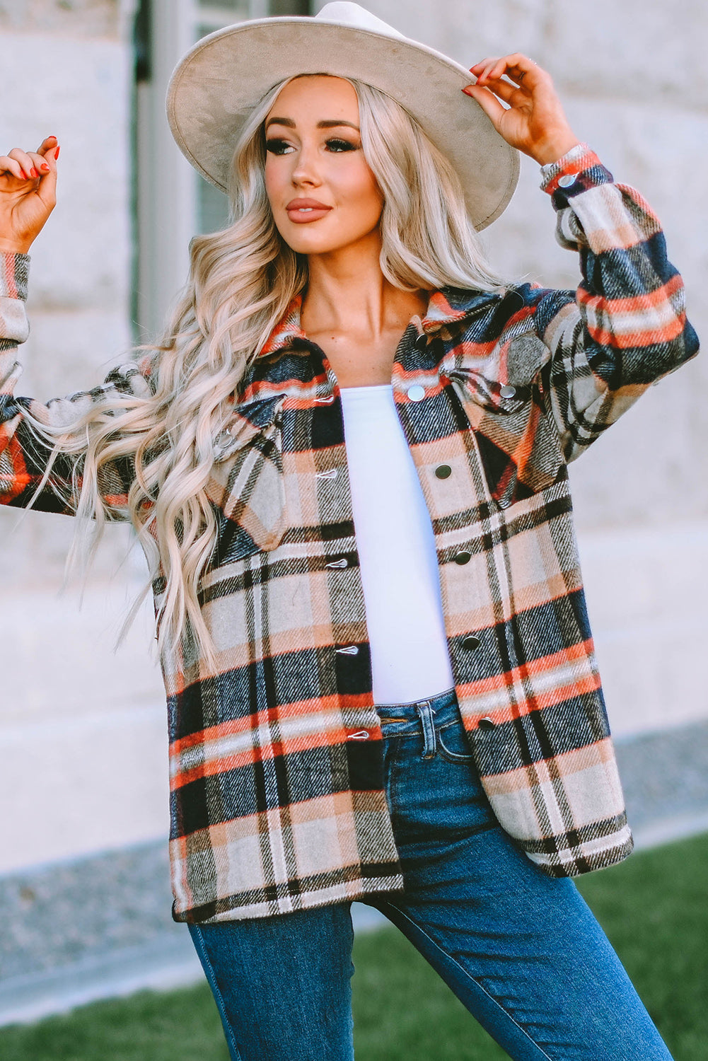 A person wearing a Brown Plaid Button Up Long Sleeve Flannel Shacket layered over a white top with ripped jeans and holding a brown handbag stands against a textured gray wall.