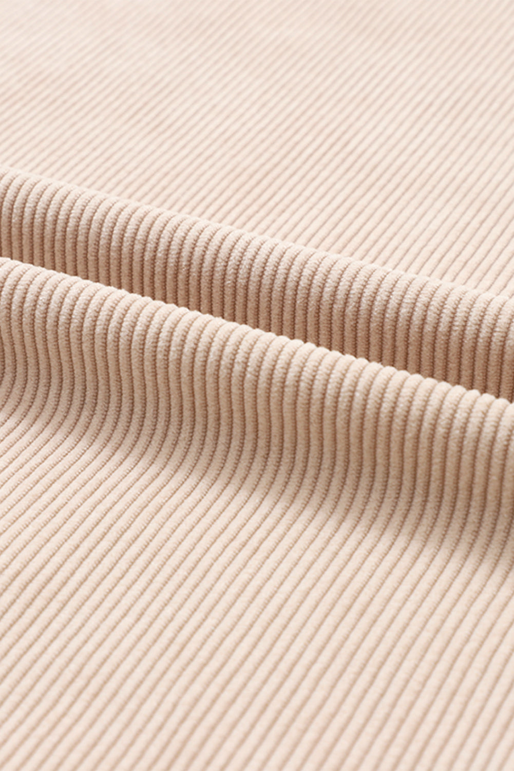 A woman stands outdoors, looking down while touching her hair with one hand. She is wearing an Apricot Ribbed Corduroy Oversized Sweatshirt and white pants, exuding an effortlessly chic and fashionable look.