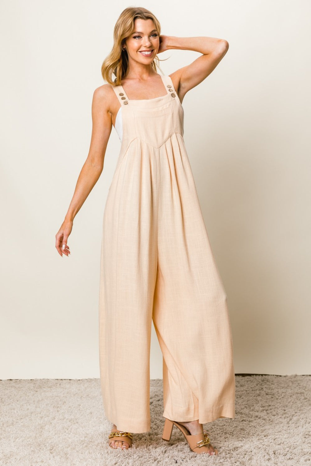 A person stands on a carpeted floor, wearing the BiBi Texture Sleeveless Wide Leg Jumpsuit in cream and beige high-heeled shoes.