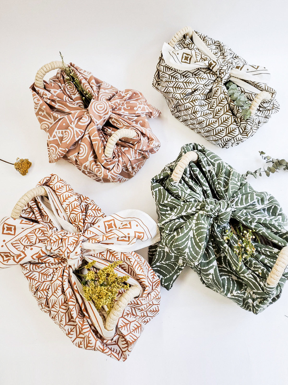 A decorative ceramic plate is beautifully set in a handwoven seagrass basket featuring handles, called the Bread Warmer & Basket Gift Set with Tea Towel - Flower. Below the basket are two patterned towels, crafted by Fair Trade artisans, showcasing leaf designs in black and orange.