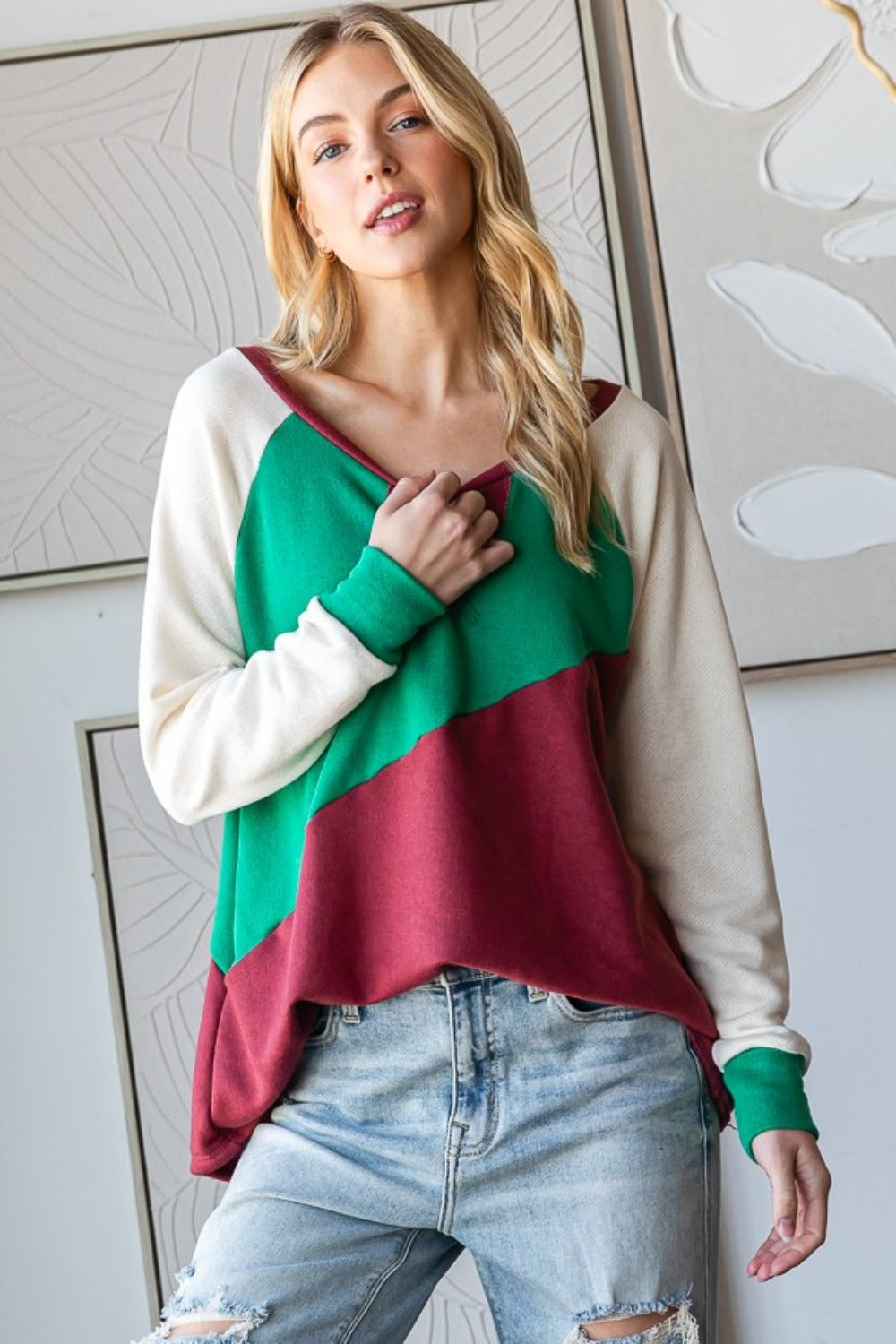 A person with long, wavy hair is wearing a HOPELY Full Size Color Block Long Sleeve T-Shirt in green, red, and white paired with light blue jeans, standing indoors against a patterned wall.