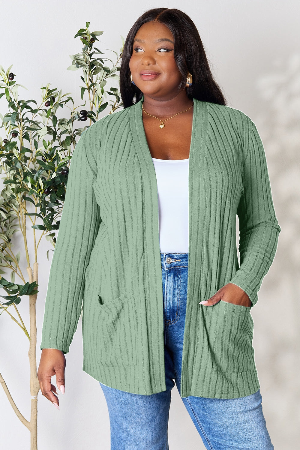 A woman in a Basic Bae Full Size Ribbed Open Front Cardigan with Pockets and jeans, made from stretchy material, stands in front of a plant, smiling slightly and looking to the side.