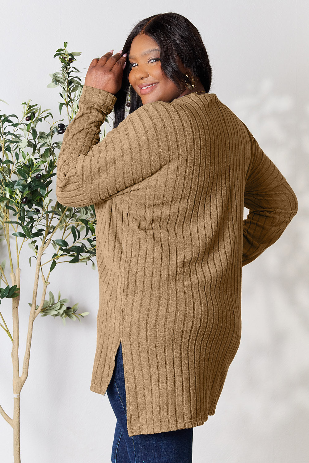 A person with shoulder-length hair smiles while wearing a beige ribbed sweater called the Basic Bae Full Size Ribbed Round Neck Long Sleeve Slit Top, along with matching beige pants, showcasing a basic style. They stand next to a green plant.