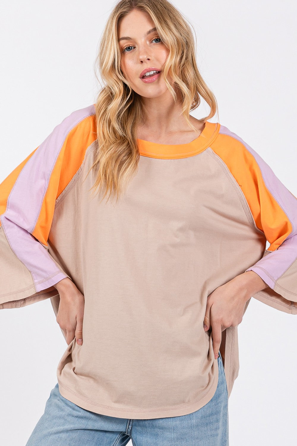 Person wearing a chic modern look with the SAGE + FIG Color Block Curved Hem T-Shirt in beige, featuring orange and purple accents, paired with blue jeans and a hat, poses against a plain background.