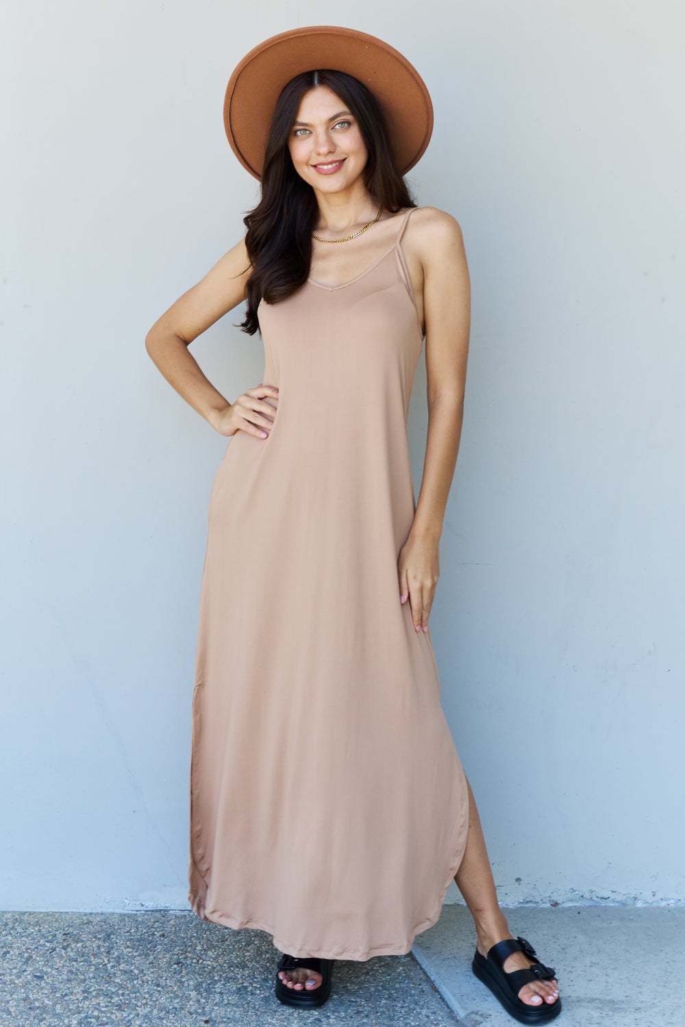 A woman stands against a plain background, wearing the Ninexis Good Energy Full Size Cami Side Slit Maxi Dress in Camel along with a wide-brimmed hat. She has one hand on her hip and is smiling.