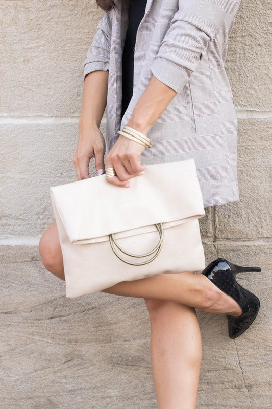 A person holds a tan Fold-over O-ring Clutch with a circular handle in front of their floral skirt and cream top.