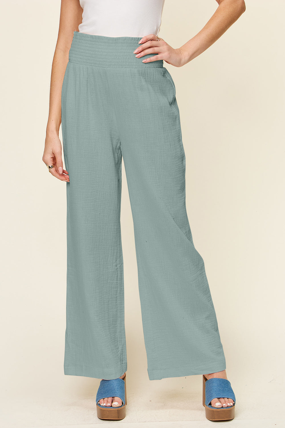 A person wearing Double Take Full Size Texture Smocked Waist Wide Leg Pants in black and a white 100% cotton top is standing with one hand on their hip. They are also wearing blue open-toe sandals.