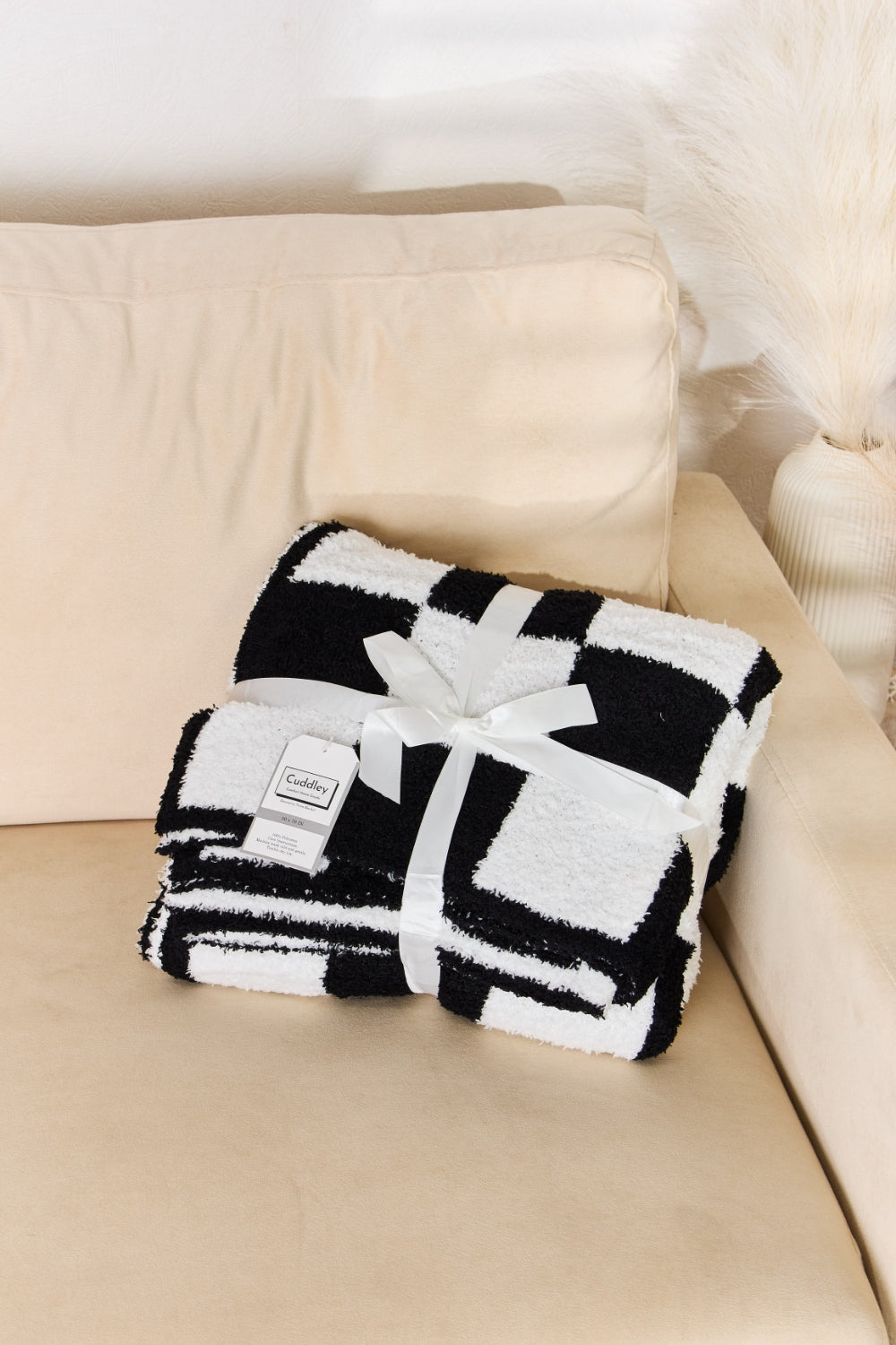 A person stands holding a large Cuddley Checkered Decorative Throw Blanket, smiling, in a room with a sofa and a potted plant. The blanket features a black and white checkerboard pattern, is made from 100% polyester, and imported for quality.