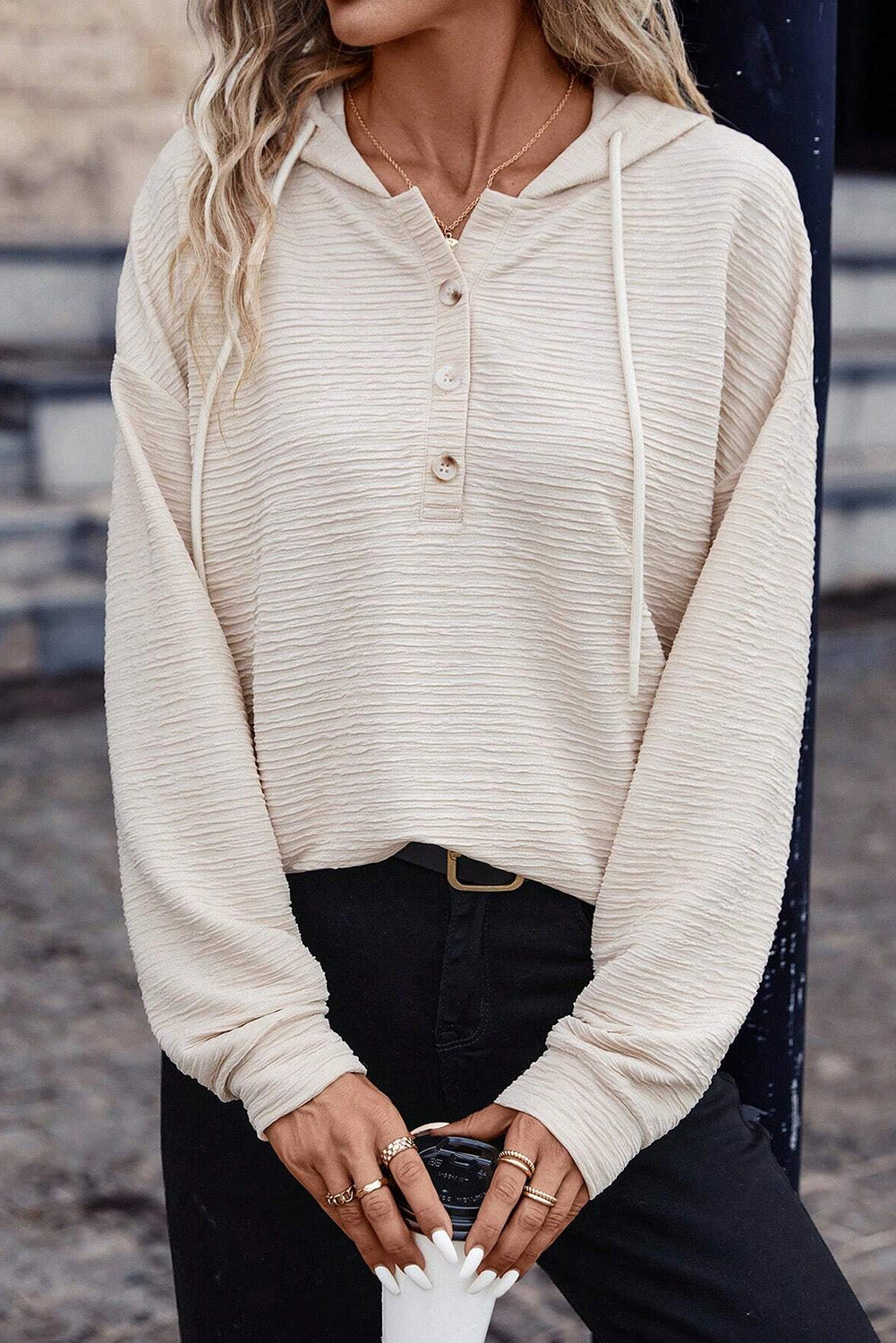 A person is wearing an Apricot Solid Color Textured Drawstring Henley Hoodie and black pants. They are holding a beverage cup and have rings on their fingers. Their face is not visible, making it perfect for casual occasions.