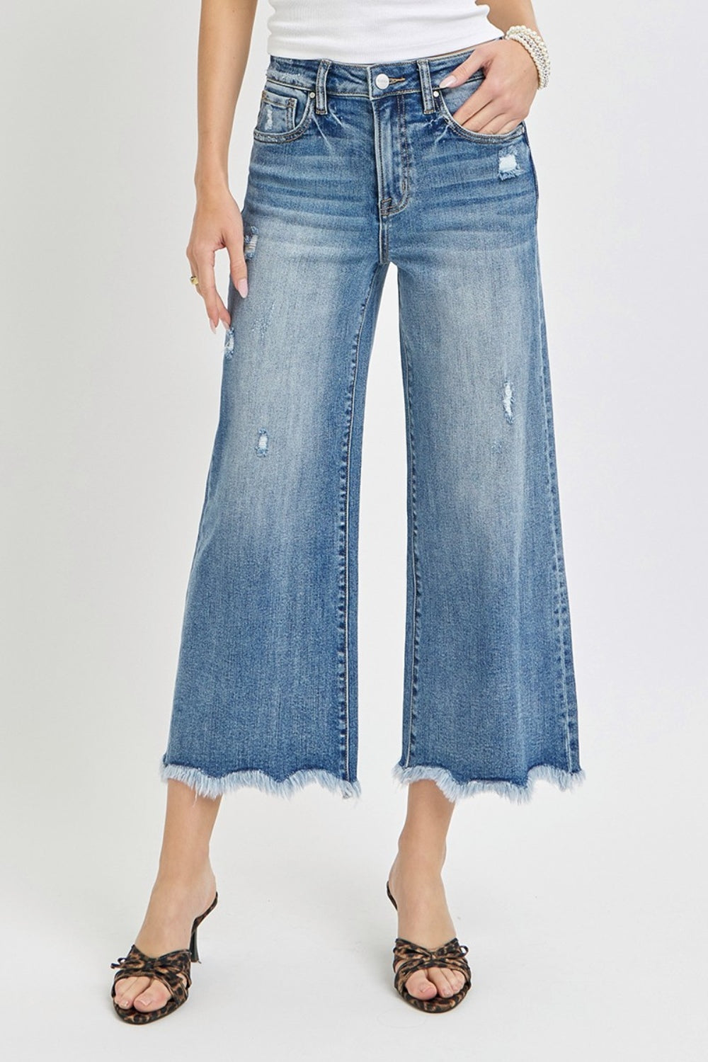 Person wearing Risen High Rise Cropped Flare Jeans with frayed details and a few rips, paired with strappy high-heeled sandals against a plain background.