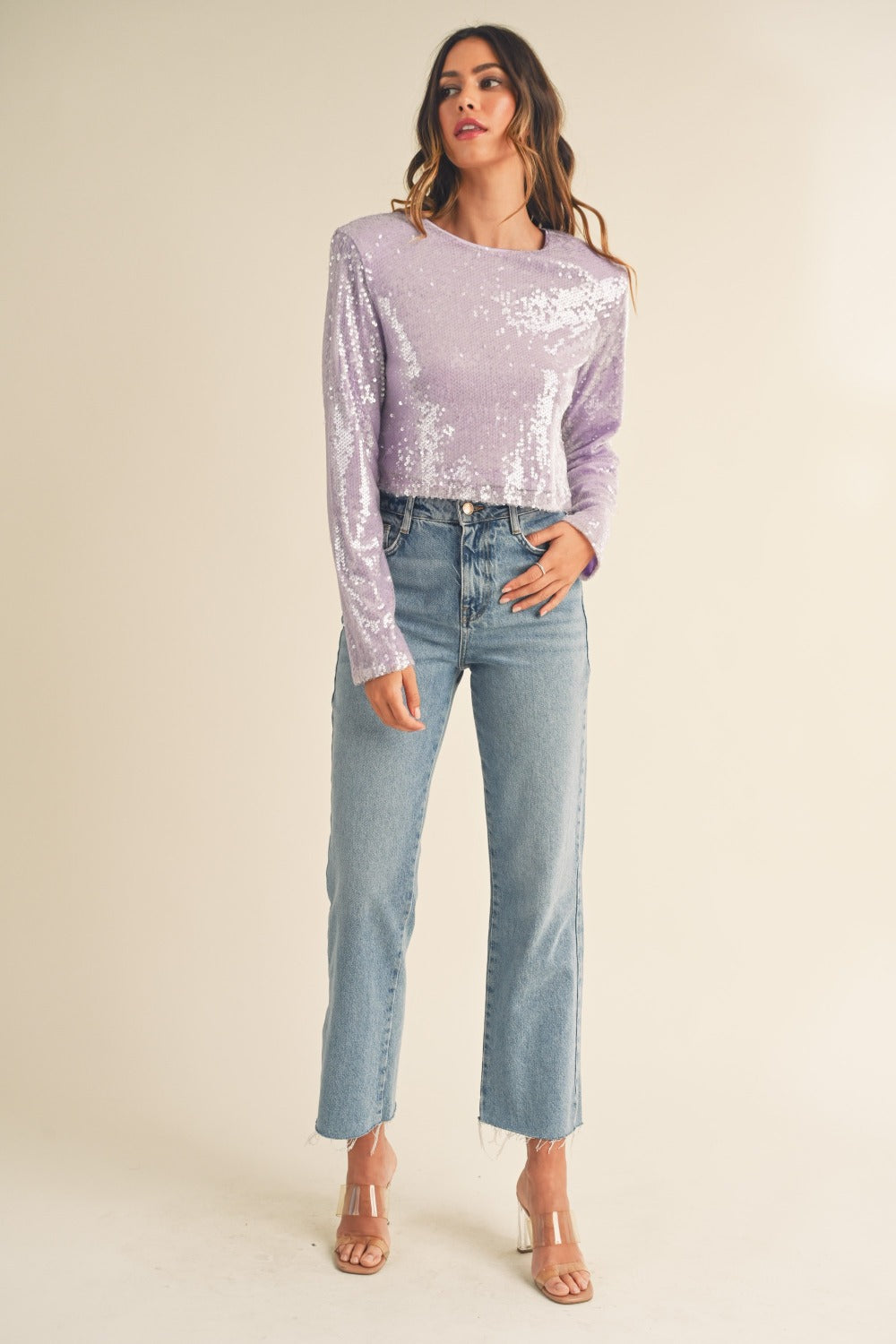 A woman with long, wavy hair wears a trendy MABLE Shoulder Padded Sequin Crop Top in light purple along with light blue jeans, standing against a plain off-white background.