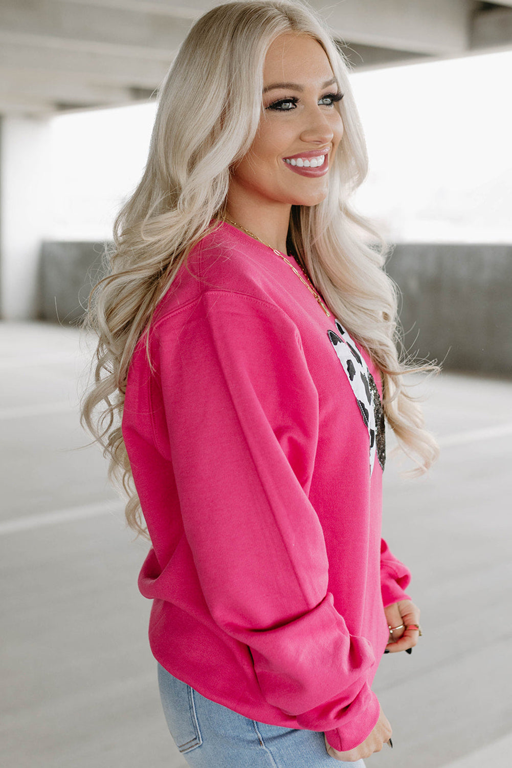 A person with long blonde hair, seen from the back, is wearing a Strawberry Pink Cow & Sequin Double Heart Patch Graphic Sweatshirt and light blue jeans.