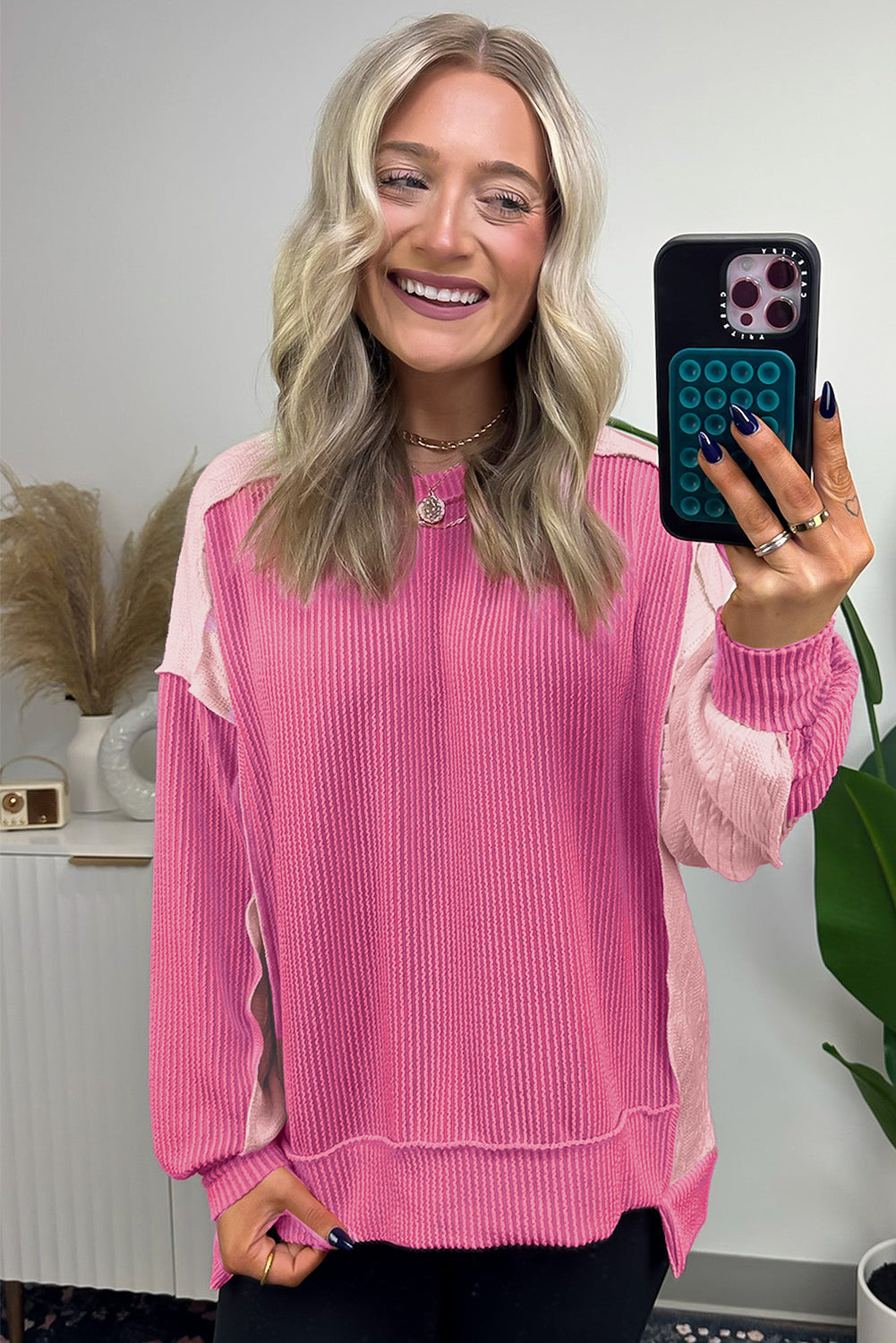 A person with long, wavy blonde hair is taking a selfie in a mirror, wearing the Bonbon Cable Knit Colorblock Exposed Seam Sweatshirt. A plant and decor items are visible in the background.