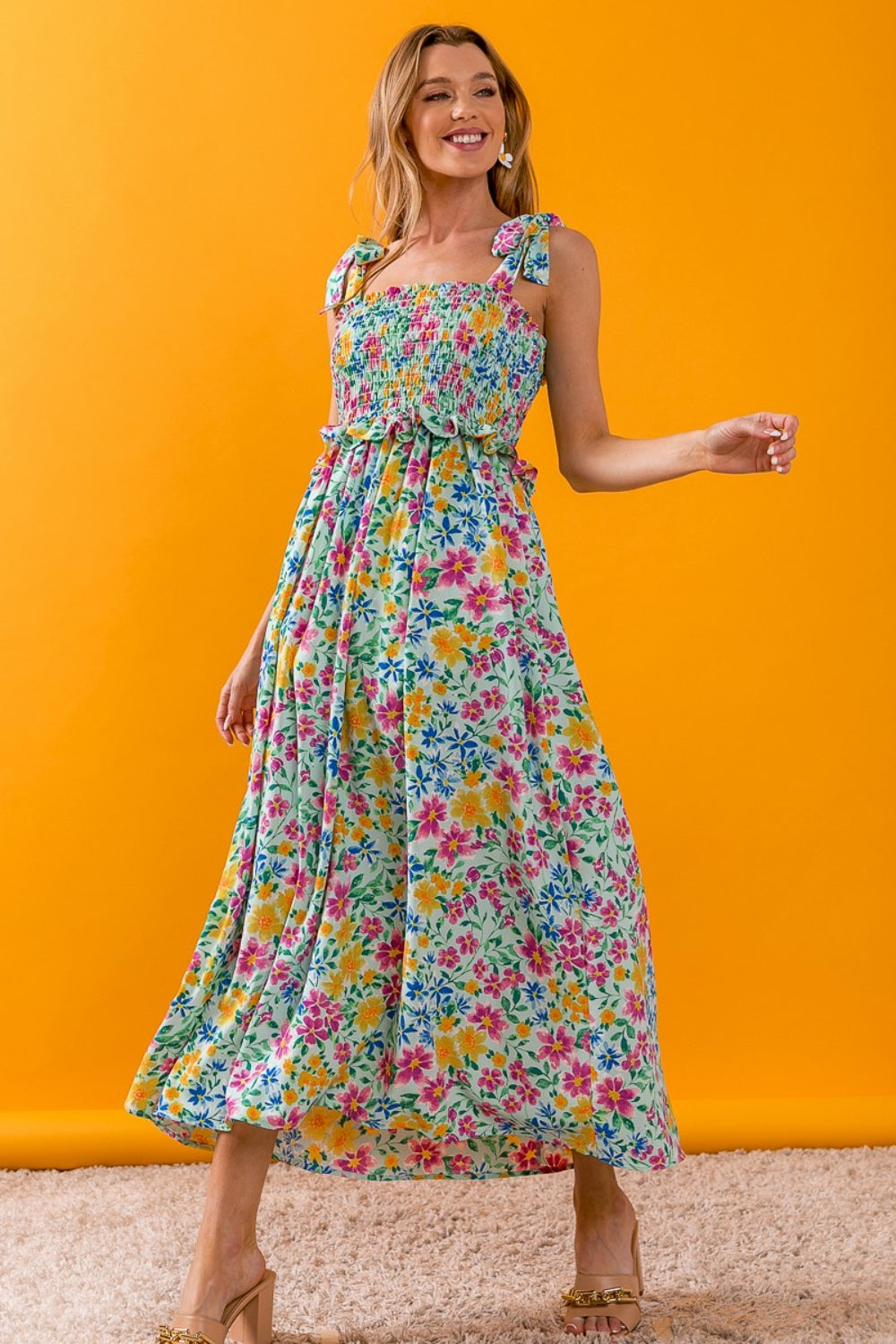 A person wearing the BiBi Floral Ruffle Trim Smocked Cami Dress with beige sandals stands on a beige carpet, posing against a white backdrop.