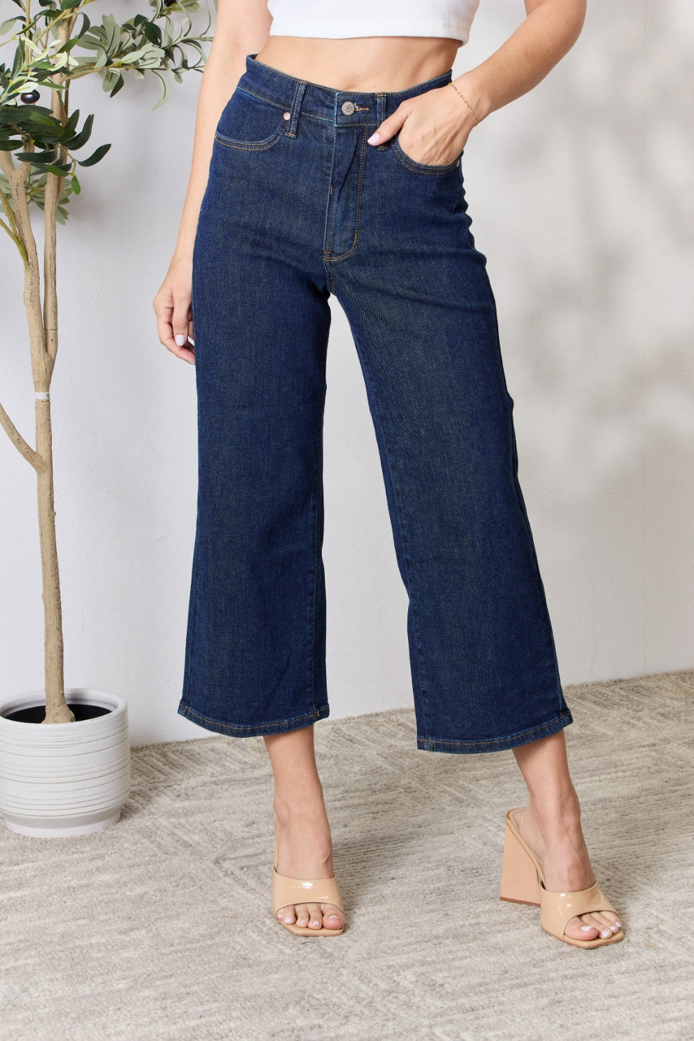 A person wearing the Judy Blue Full Size High Waist Cropped Wide Leg Jeans, which are renowned for their tummy control design, and nude block heel sandals stands near a potted plant.