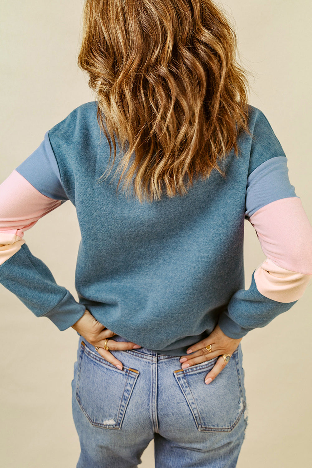 A rear view of a person with wavy hair, dressed in a Blue Colorblock Long Sleeve Pullover Sweatshirt and light blue jeans, hands resting on their hips. Perfect for winter fashion, this versatile piece is an ideal addition to any wardrobe.