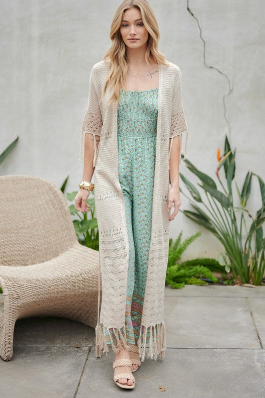 A woman in a green floral dress and a Solid Long Cardigan With Fringe exudes boho-chic vibes as she strolls on a patio, surrounded by outdoor furniture and lush plants.