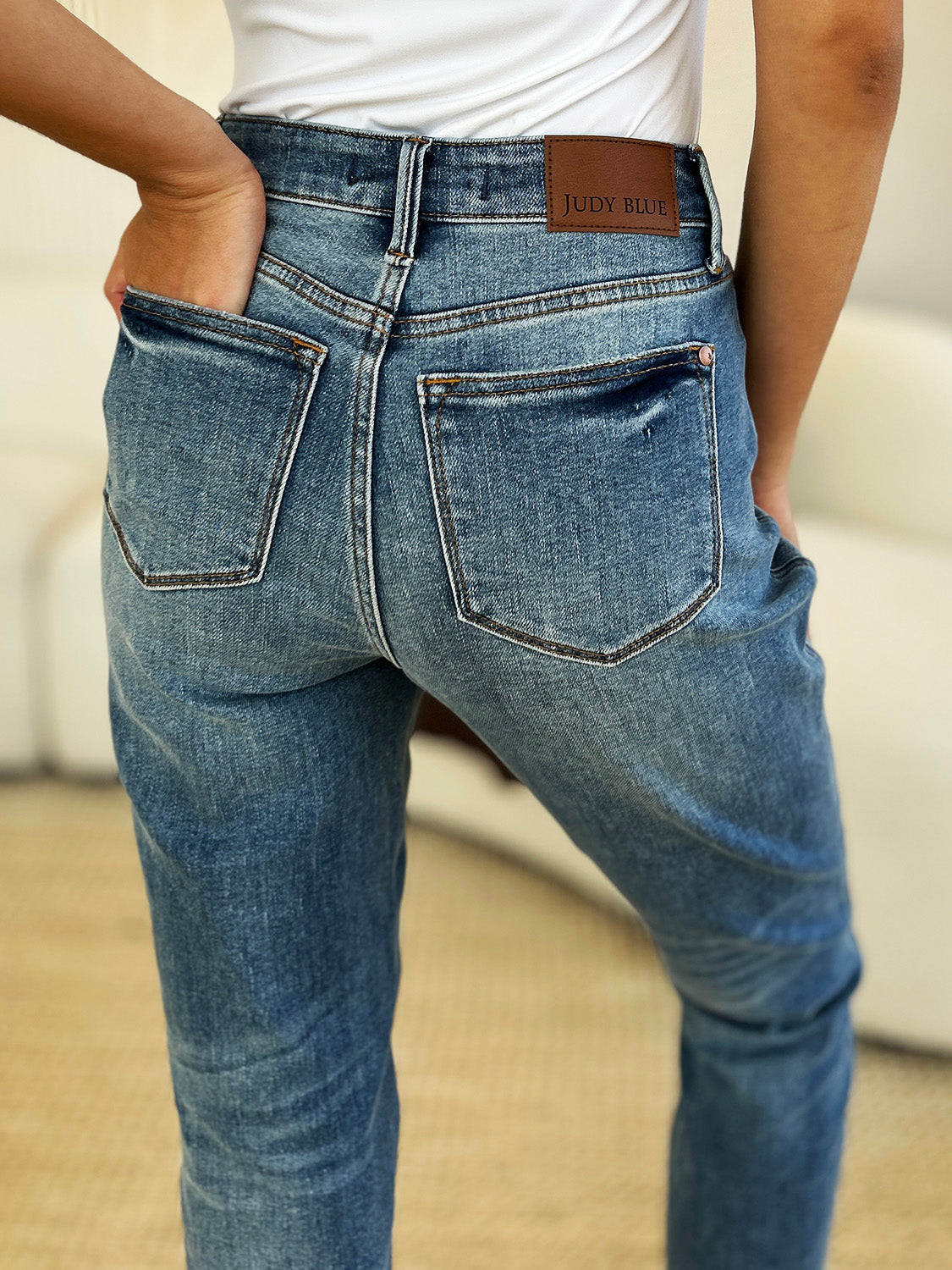 Person wearing a black top, Judy Blue Full Size Mid Rise Rigid Magic Release Hem Jeans, and black sneakers with white laces, standing on a beige carpet with hands in pockets.