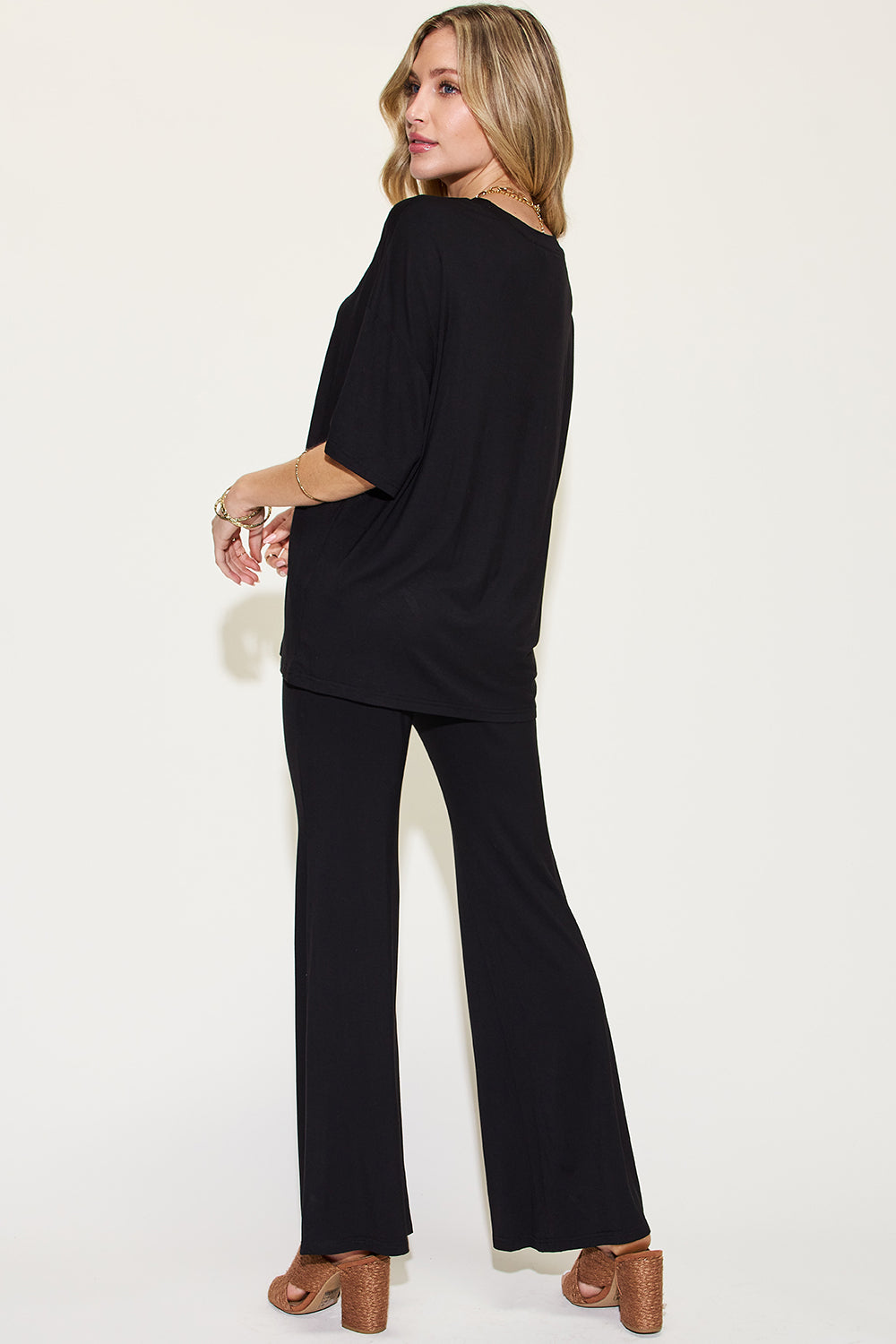 A woman with long, blonde hair wears the Basic Bae Full Size Bamboo Drop Shoulder T-Shirt and Flare Pants Set in black, paired with brown heeled sandals and layered necklaces. She stands against a plain, light background.