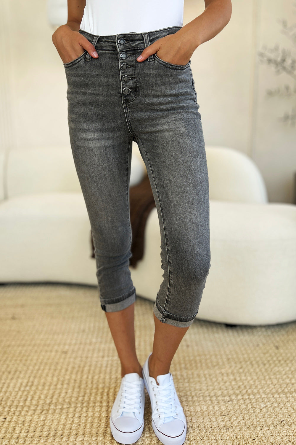 A person wearing Judy Blue Full Size Button Fly High Waist Cuffed Capris in gray, paired with a white top and white sneakers, stands on a beige carpeted floor.