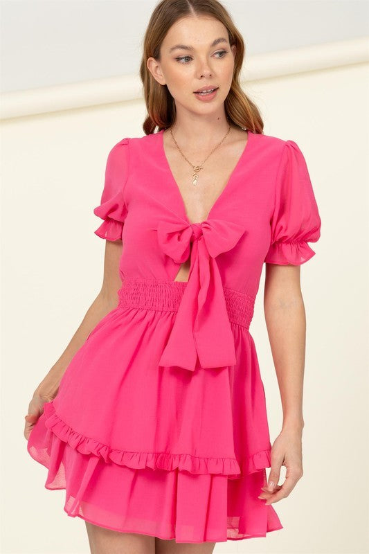 A woman is wearing the "Days Like This Tie-Front Mini Dress" in a vibrant pink color, featuring short ruffled sleeves, a prominent bow at the chest, and a tiered skirt with a smocked waist.