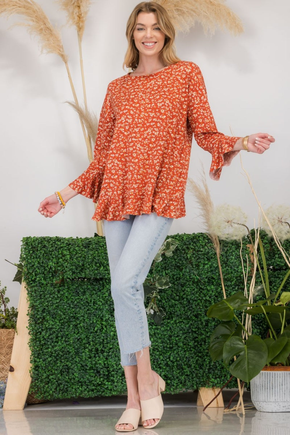 A woman wearing the Celeste Full Size Floral Ruffle Detail Top and jeans stands smiling in front of pampas grass and greenery, exuding feminine flair.