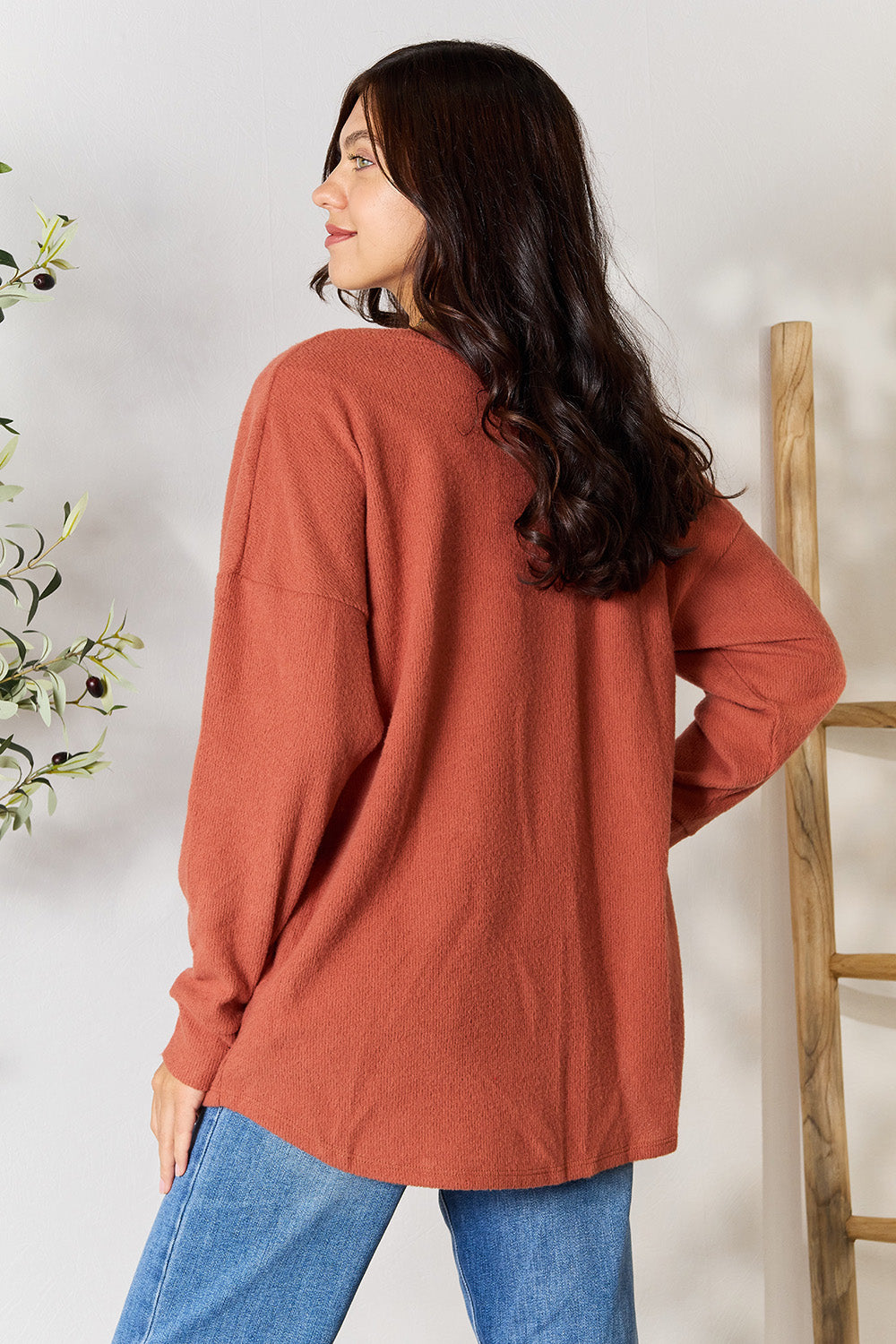 A person wearing the BOMBOM Drop Shoulder Long Sleeve Blouse with Pockets, featuring a cozy, rust color and relaxed fit, stands against a plain wall with a plant in the background.