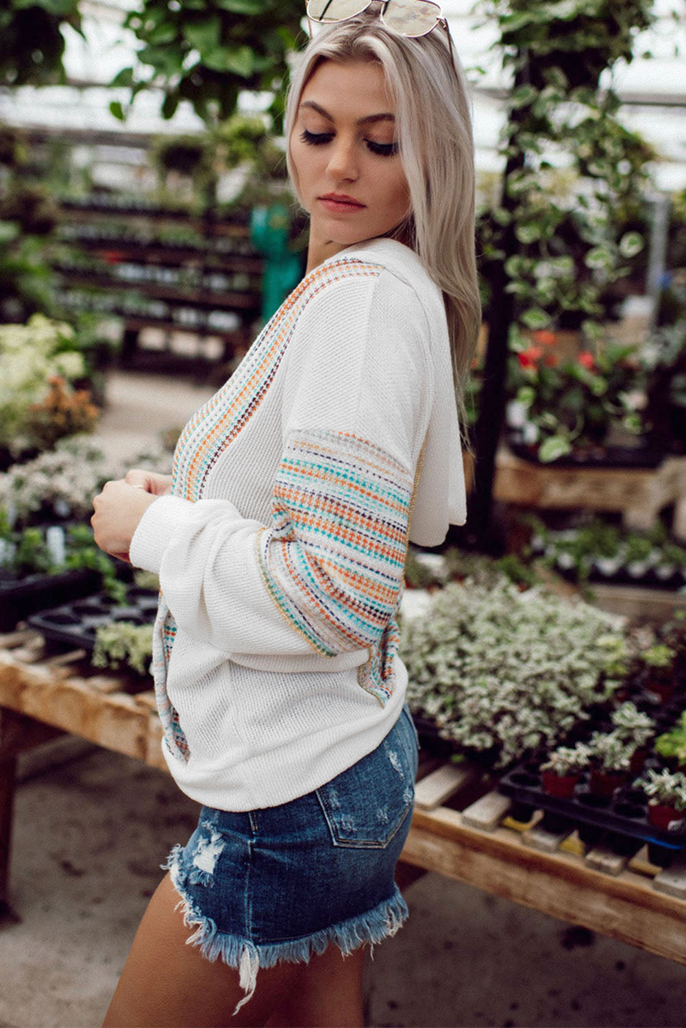 A woman with long blonde hair wearing a Multicolor Striped Patchwork V Neck Drop Shoulder Knit Hoodie and denim shorts stands in a plant nursery. She has sunglasses perched on her head and is looking down.