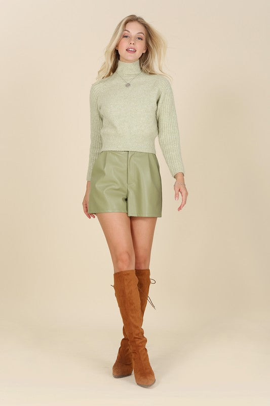 An individual is dressed in a light green sweater paired with pleated vegan leather shorts, featuring a functional pocket where one hand rests.