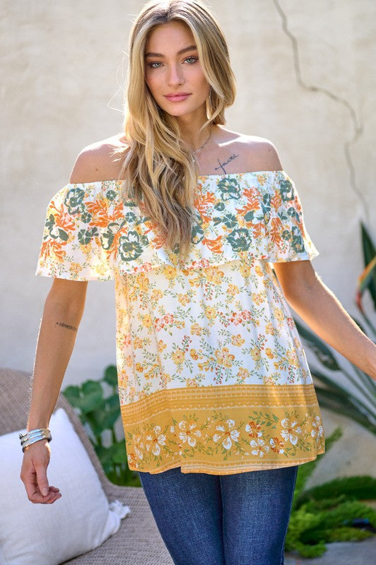 A woman with blonde hair is smiling while standing outdoors, wearing the Printed Off Shoulder Smocked Top paired with jeans.