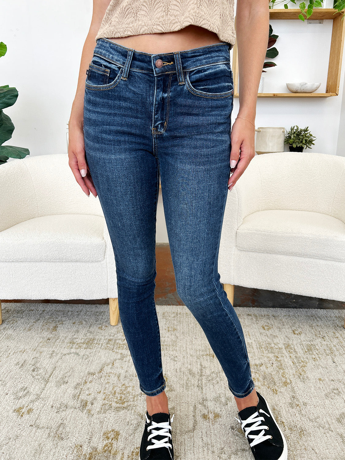 A person stands indoors wearing Judy Blue Full Size Classic Handsand Skinny Jeans, a beige top, and black sneakers with white laces. Their right hand is in their pocket. A partial view of two white chairs is visible in the background. The jeans feature a hand-sanded finish for a flattering fit.