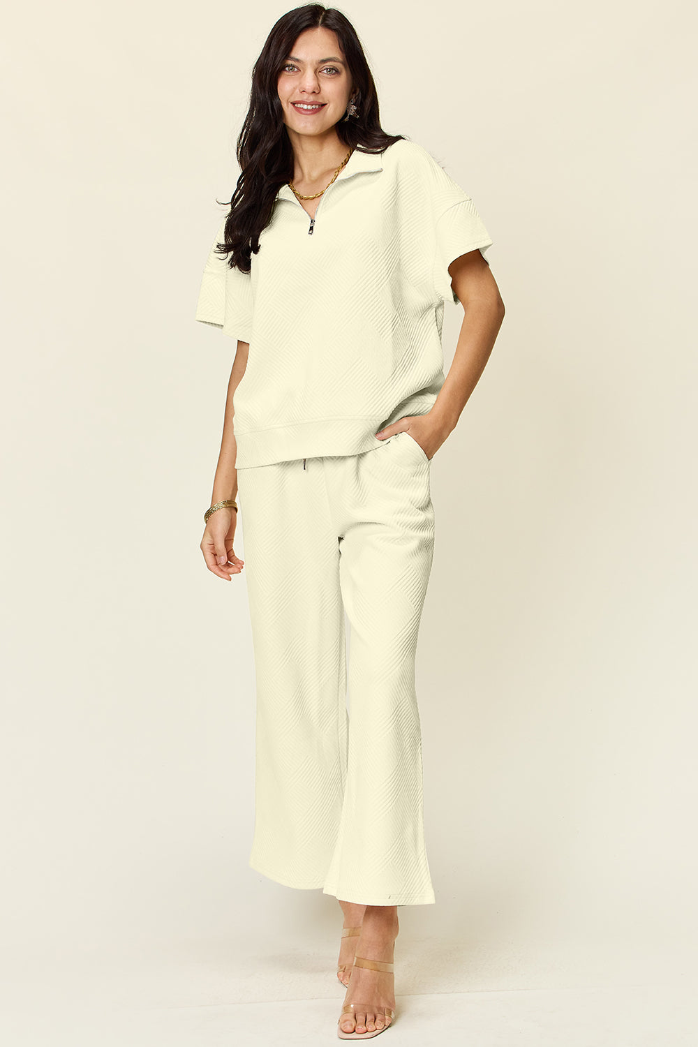 A woman is wearing the Double Take Full Size Texture Half Zip Short Sleeve Top and Pants Set, featuring a white zip-up short-sleeve top and matching wide-leg, drawstring pants. She stands with one hand in her pocket and smiles against a light background.