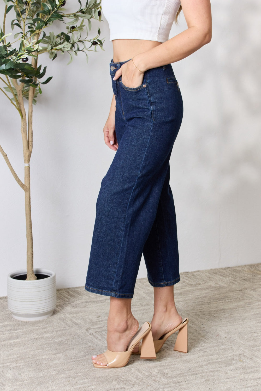 A person wearing the Judy Blue Full Size High Waist Cropped Wide Leg Jeans, which are renowned for their tummy control design, and nude block heel sandals stands near a potted plant.